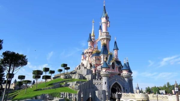 Une image vibrante du château de la Belle au Bois Dormant à Disneyland Paris, avec des flèches roses et bleues sous un ciel bleu clair avec de la verdure entourant la base.