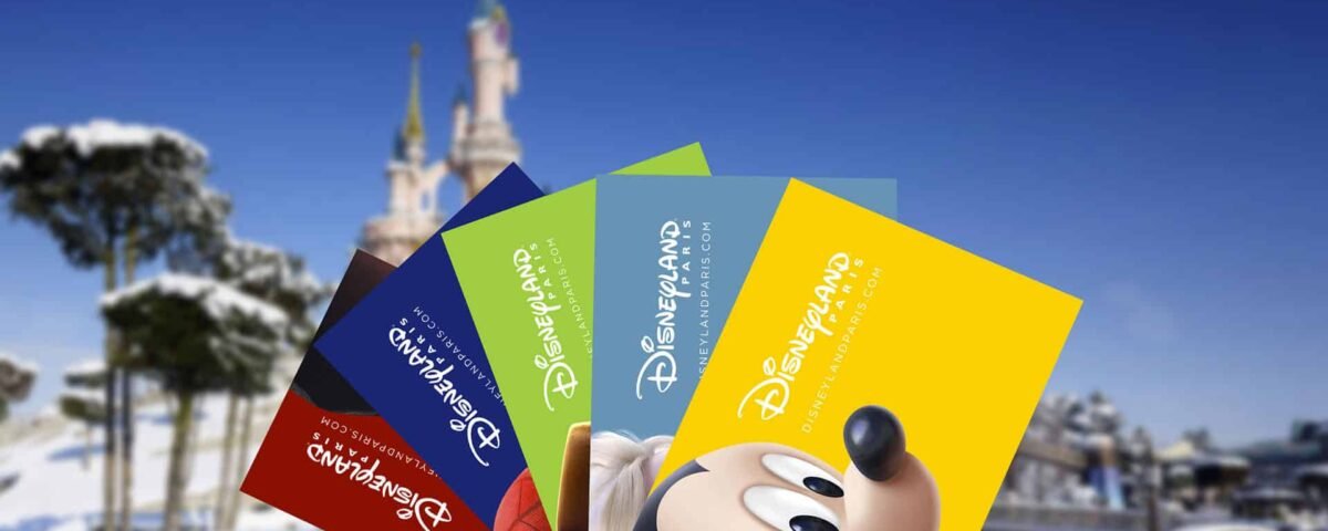 Une collection de billets colorés de Disneyland Paris représentant Mickey Mouse, affichés au premier plan avec un château Disneyland enneigé en arrière-plan sous un ciel bleu clair.