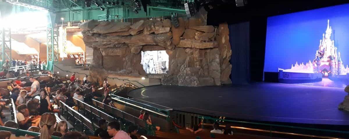 Public assis dans un théâtre doté d’une grande scène présentant un décor rocheux et un décor de Disneyland en toile de fond. Au-dessus de nous, de nombreux éclairages de scène et équipements de gréage sont visibles.