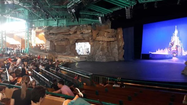 Public assis dans un théâtre doté d’une grande scène présentant un décor rocheux et un décor de Disneyland en toile de fond. Au-dessus de nous, de nombreux éclairages de scène et équipements de gréage sont visibles.