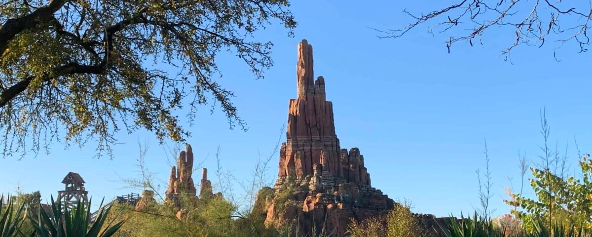 Une grande formation rocheuse et désertique se dressant bien en évidence sous un ciel bleu clair, ressemblant au décor sur le thème de la frontière de Big Thunder Mountain à Disneyland Paris, entourée d'un feuillage vert luxuriant et de plantes de cactus dans