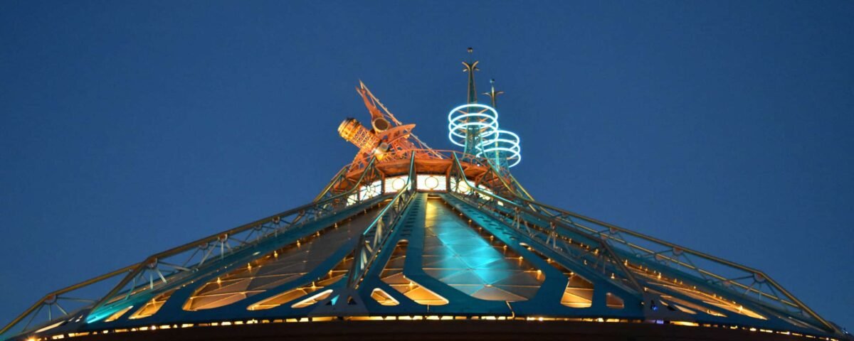 Une photo nocturne vibrante d'un manège "De la Terre à la Lune" à Disneyland Paris, mettant en vedette une structure en forme de moulin à vent illuminée de chaudes lumières orange et turquoise, sur un fond sombre