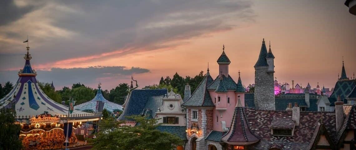 Une vue panoramique d'un parc d'attractions sur le thème de Disneyland au crépuscule, présentant une architecture fantaisiste avec des tourelles et un carrousel sous un ciel coloré.