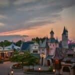 Une vue panoramique d'un parc d'attractions sur le thème de Disneyland au crépuscule, présentant une architecture fantaisiste avec des tourelles et un carrousel sous un ciel coloré.