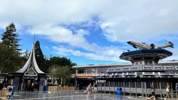 Une scène vibrante à Disneyland Paris à Discoveryland avec un manège futuriste sur le thème de l'espace, Star Tours, avec un grand modèle de vaisseau spatial sur le dessus. Les gens se promènent sous un ciel bleu tacheté de nuages, près de l'humidité réfléchissante