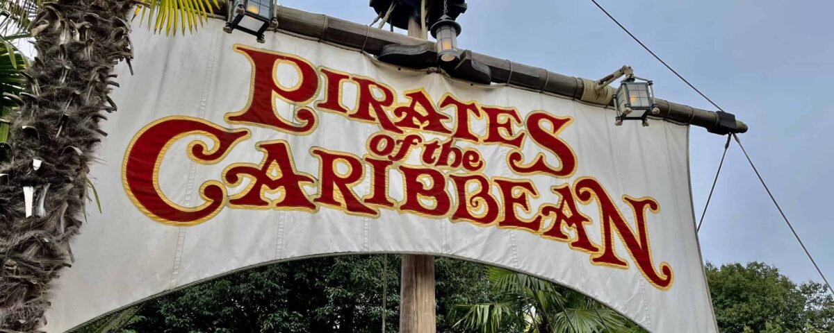 Un panneau indiquant « pirates des caraïbes » en lettres rouges et jaunes stylisées et audacieuses, accroché à une entrée flanquée de verdure luxuriante et de palmiers.