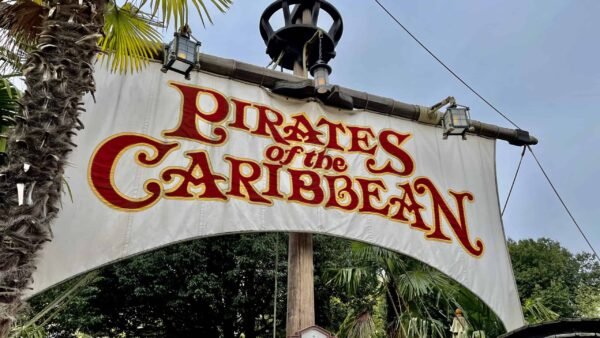 Un panneau indiquant « pirates des caraïbes » en lettres rouges et jaunes stylisées et audacieuses, accroché à une entrée flanquée de verdure luxuriante et de palmiers.