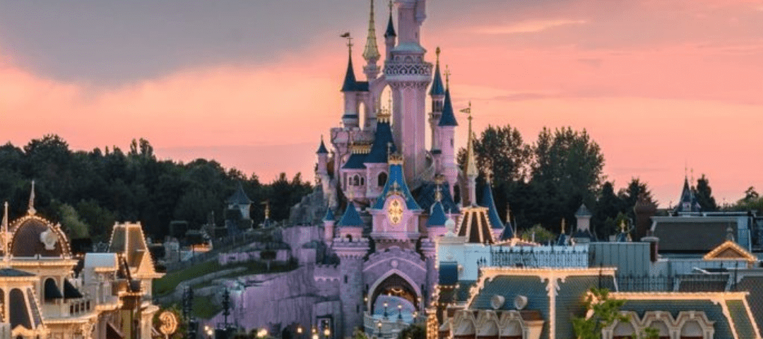 Une vue pittoresque de Disneyland Paris avec le château emblématique au centre, éclairé sous un magnifique coucher de soleil rose et orange, avec des foules de visiteurs en contrebas.