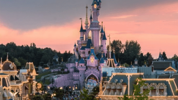 Une vue pittoresque de Disneyland Paris avec le château emblématique au centre, éclairé sous un magnifique coucher de soleil rose et orange, avec des foules de visiteurs en contrebas.