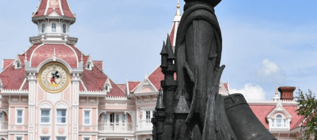 Statue de style silhouette de Mickey Mouse tenant une baguette, avec en arrière-plan la façade rose et blanche du grand bâtiment de Disneyland Paris avec une tour d'horloge.