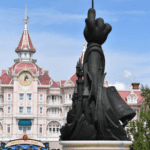Statue de style silhouette de Mickey Mouse tenant une baguette, avec en arrière-plan la façade rose et blanche du grand bâtiment de Disneyland Paris avec une tour d'horloge.