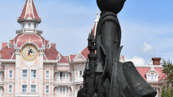Statue de style silhouette de Mickey Mouse tenant une baguette, avec en arrière-plan la façade rose et blanche du grand bâtiment de Disneyland Paris avec une tour d'horloge.