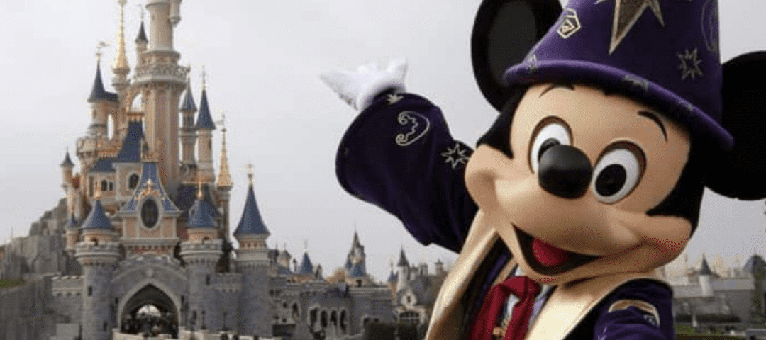 Mickey Mouse, en costume de sorcier, fait un geste vers la caméra devant le château emblématique des studios Walt Disney par une journée nuageuse.