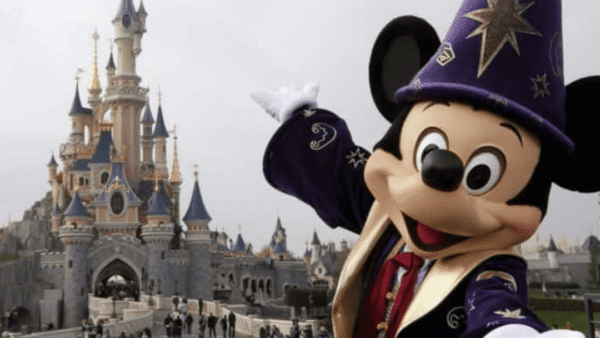 Mickey Mouse, en costume de sorcier, fait un geste vers la caméra devant le château emblématique des studios Walt Disney par une journée nuageuse.