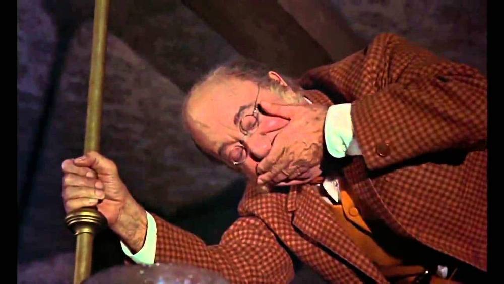 Un homme âgé avec des lunettes, vêtu d'une veste en tweed et d'un gilet, s'assoupit avec sa main couvrant sa bouche, appuyé sur une canne en bois dans une scène du film Disney.
