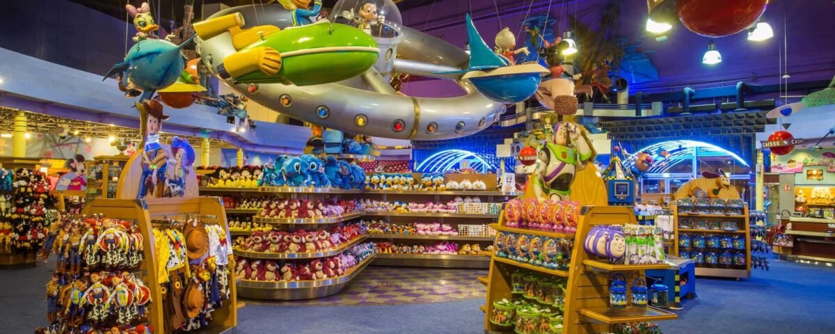 Un vaste magasin de jouets intérieur avec un présentoir de vaisseau spatial coloré suspendu au plafond au-dessus d'étagères soigneusement disposées de jouets et d'animaux en peluche, proposant un service de shopping Disneyland Paris.