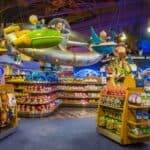 Un vaste magasin de jouets intérieur avec un présentoir de vaisseau spatial coloré suspendu au plafond au-dessus d'étagères soigneusement disposées de jouets et d'animaux en peluche, proposant un service de shopping Disneyland Paris.