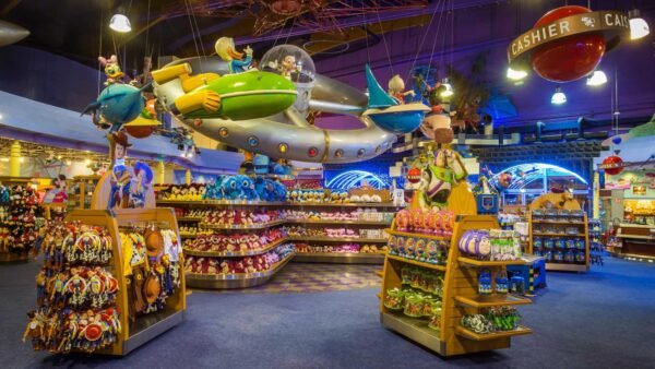 Un vaste magasin de jouets intérieur avec un présentoir de vaisseau spatial coloré suspendu au plafond au-dessus d'étagères soigneusement disposées de jouets et d'animaux en peluche, proposant un service de shopping Disneyland Paris.