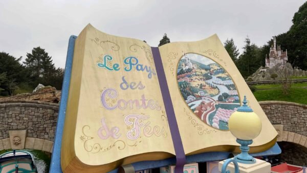 Un grand livre de contes ouvert intitulé "le pays des contes de fées" à côté d'une promenade en bateau de conte de fées, avec une peinture colorée détaillée d'une scène de village sur les pages, et Raiponce