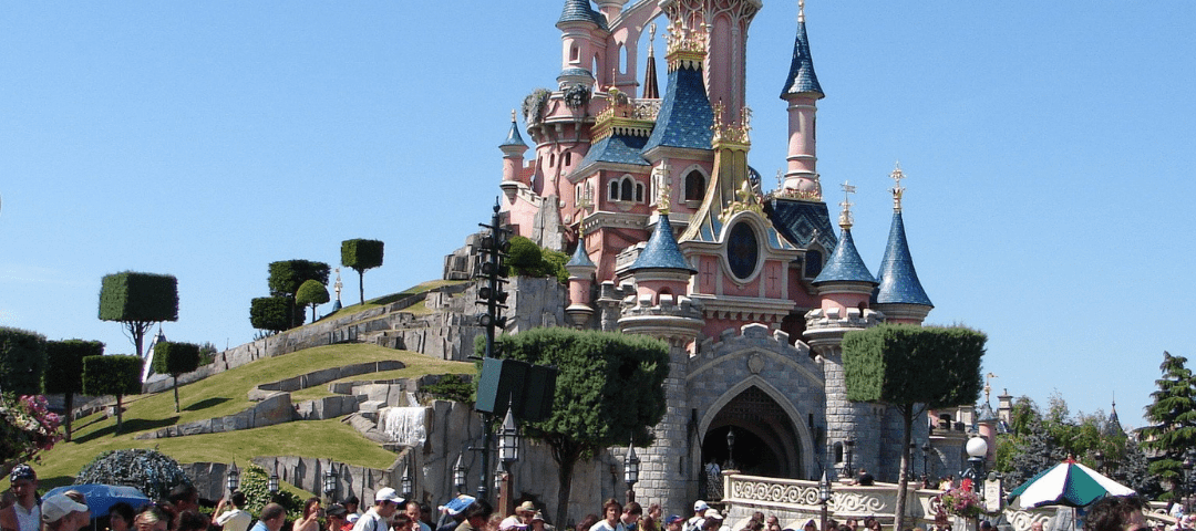 Foule de visiteurs profitant d'une journée ensoleillée à des horaires précis devant l'emblématique château de la Belle au Bois Dormant rose et bleu à Disneyland.