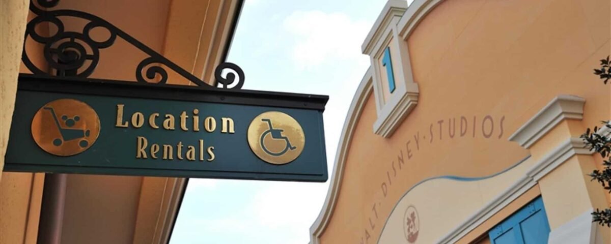 Un panneau indiquant « location d'emplacements » avec une palette d'artiste et des icônes de fauteuil roulant est suspendu à un support en fer forgé sur un bâtiment aux murs couleur pêche et avec le texte « Disneyland Paris ».
