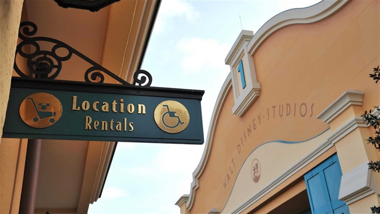 Un panneau indiquant « location d'emplacements » avec une palette d'artiste et des icônes de fauteuil roulant est suspendu à un support en fer forgé sur un bâtiment aux murs couleur pêche et avec le texte « Disneyland Paris ».
