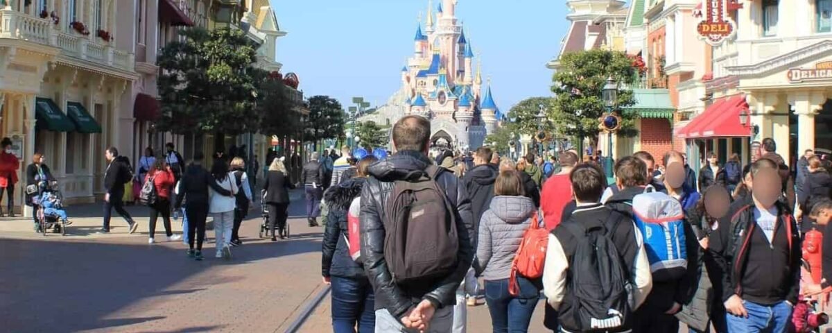 Les visiteurs connaissent une forte fréquentation en marchant dans une rue animée de Disneyland, avec l'emblématique château Disney en arrière-plan par une journée ensoleillée.