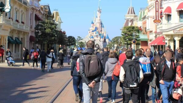 Les visiteurs connaissent une forte fréquentation en marchant dans une rue animée de Disneyland, avec l'emblématique château Disney en arrière-plan par une journée ensoleillée.
