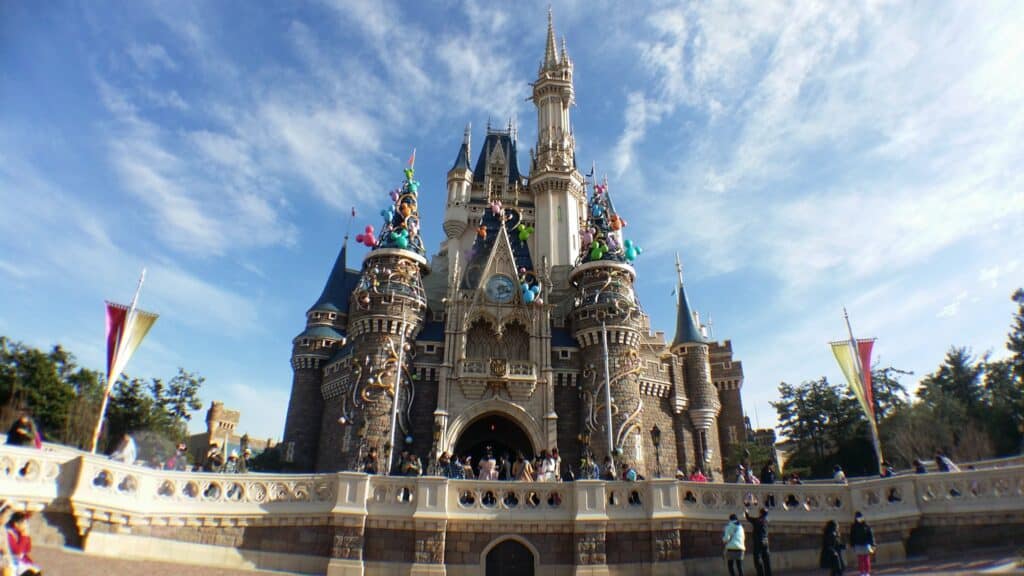 Plan large d'un château de conte de fées à Disneyland sous un ciel bleu clair, décoré de statues colorées de personnages Disney sur ses flèches et entouré d'arbres verts luxuriants.