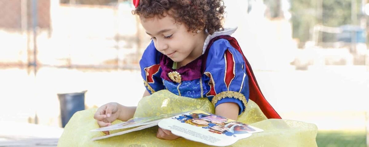 Un jeune enfant aux cheveux bouclés, vêtu d'un costume de blanche-neige jaune et bleu, lit un livre en plein air par une journée ensoleillée.