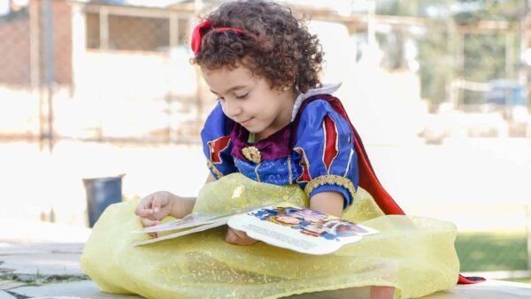Un jeune enfant aux cheveux bouclés, vêtu d'un costume de blanche-neige jaune et bleu, lit un livre en plein air par une journée ensoleillée.