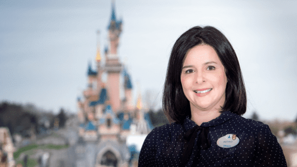 Une femme aux cheveux noirs mi-longs, vêtue d'une blouse à pois et d'un badge portant l'inscription "astrid gomez", sourit devant l'emblématique Château de la Belle au Bois Dormant de Disneyland Paris.