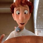 Un personnage animé, un jeune chef aux cheveux roux, sourit en tenant une cuillère en bois au-dessus d'une grande marmite de ratatouille, avec un petit rat bleu qui sort de sa veste de chef