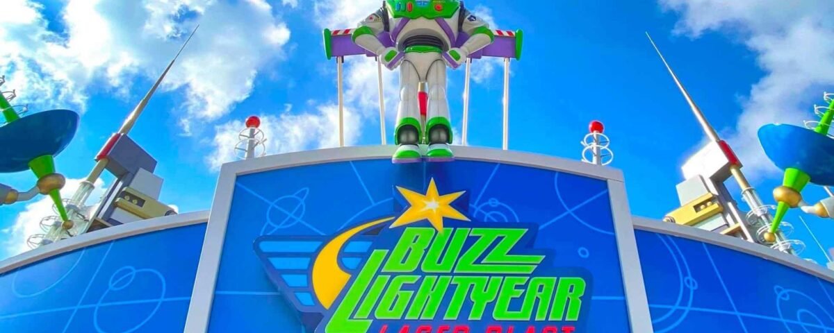 La statue de Buzz l'Éclair se dresse au sommet de l'entrée du manège Buzz l'Éclair Laser Blast sous un ciel bleu vif avec des nuages épars, entourée de décorations colorées sur le thème de l'espace, dont un « trouvez l'intr