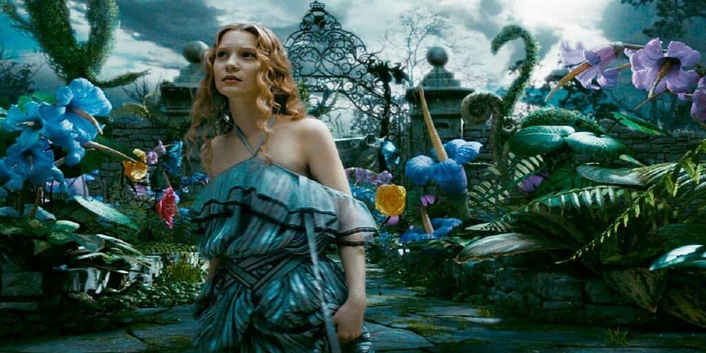 Alice, une jeune femme vêtue d'une robe fluide bleue et grise, se tient dans un jardin fantastique et vibrant avec des fleurs colorées surdimensionnées et une porte en métal ornée en arrière-plan, rappelant une scène.