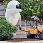 Un grand modèle du robot Eve du film "wall-e" debout à côté d'un modèle tout aussi grand de wall-e lui-même, entouré d'arbustes et adossé à un bâtiment.