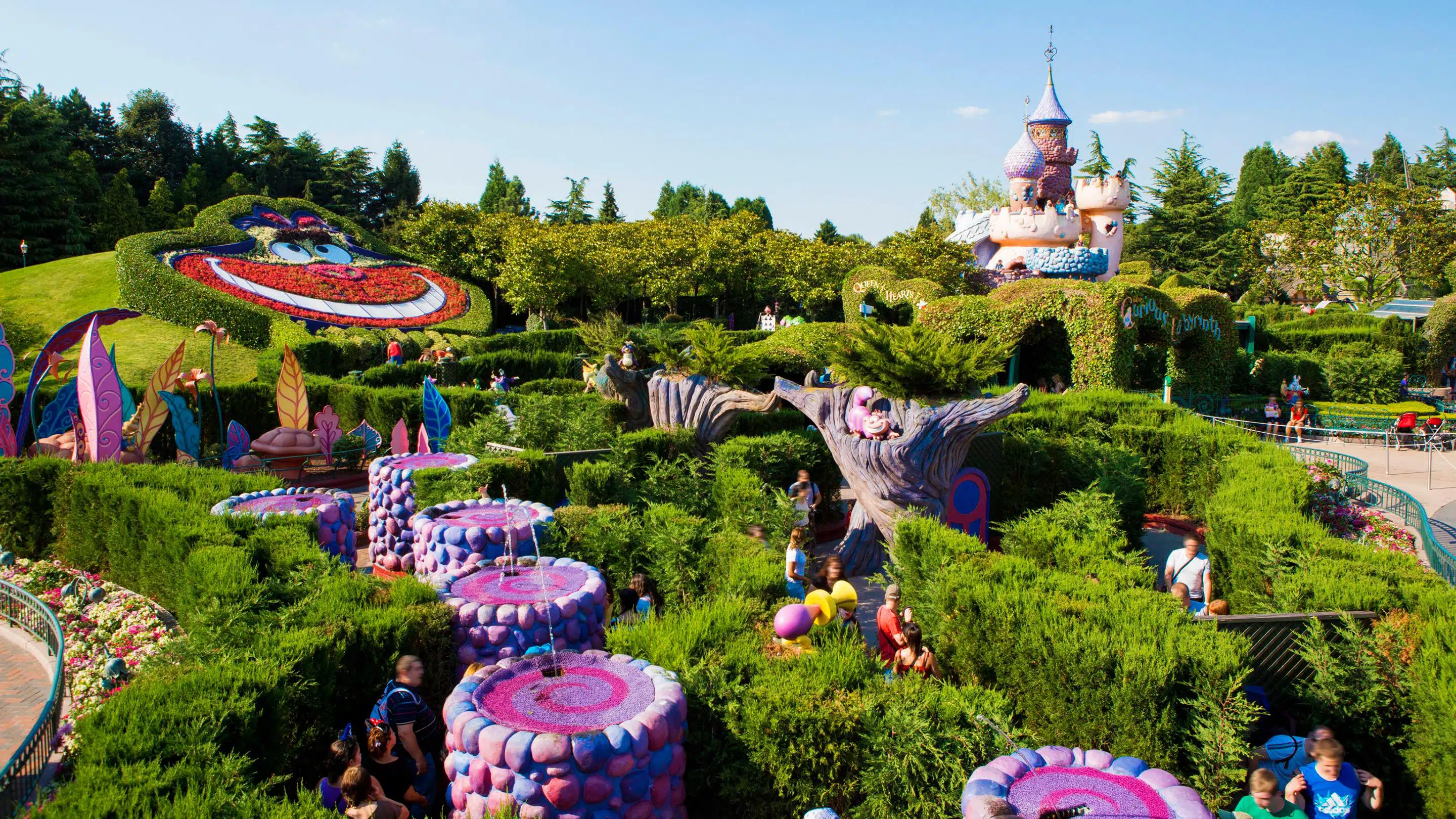 Parc à thème animé avec un jardin fantaisiste d'Alice au Pays des Merveilles avec des personnages topiaires, des compositions végétales colorées et un château de conte de fées en arrière-plan sous un ciel bleu clair.