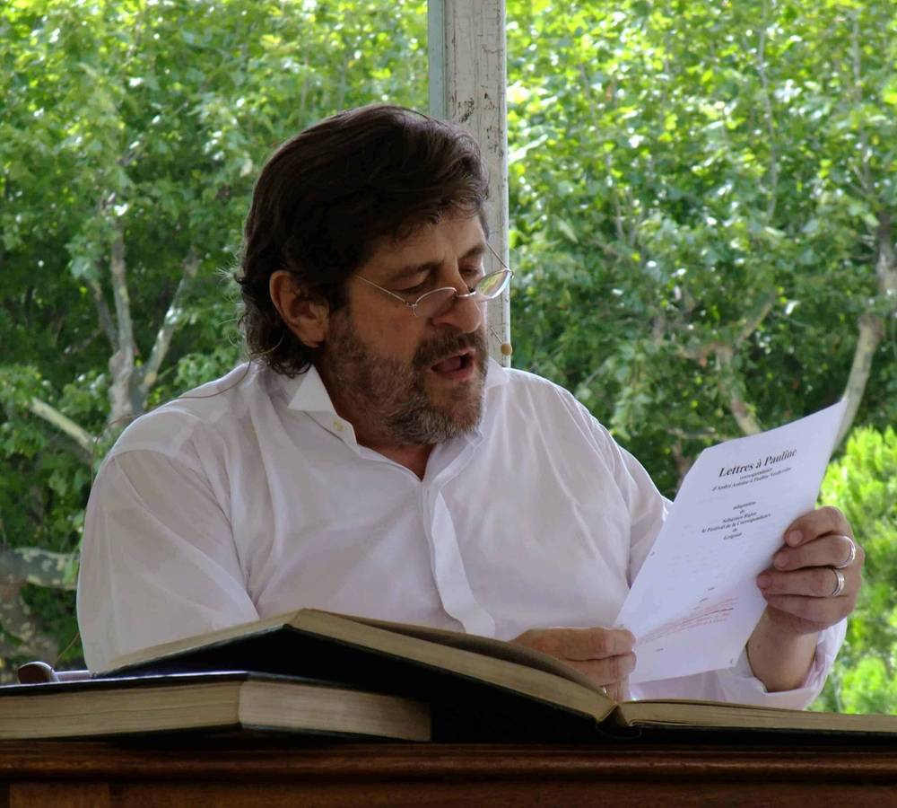 Un homme avec une barbe et des lunettes lisant un scénario assis à un bureau avec des livres ouverts, à l'extérieur avec des arbres en arrière-plan, éventuellement impliqué dans des services de doublage de films ou d'émissions de télévision.