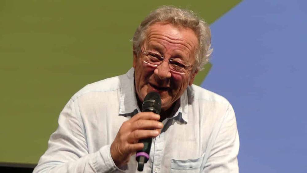 Un homme âgé aux cheveux gris et aux lunettes, souriant alors qu'il effectue un doublage dans un microphone, vêtu d'une chemise bleu clair, sur fond bleu et vert.