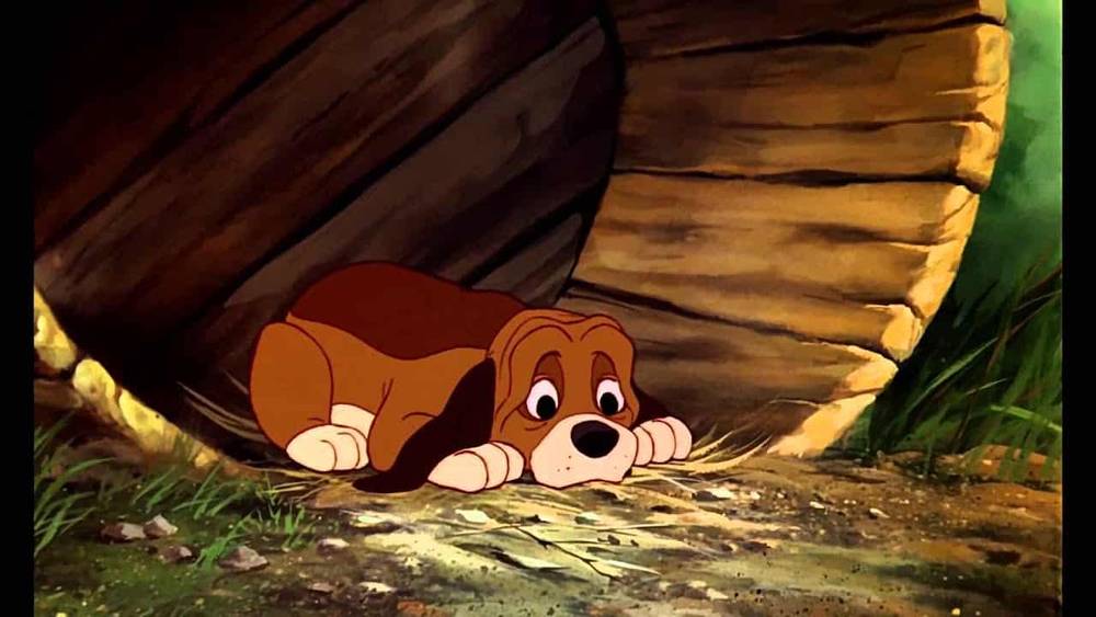 Un chiot animé marron et feu du film classique de Disney "Rox et Rouky", l'air effrayé et allongé à plat sur le ventre sous un objet en bois, regardant avec de grands yeux expressifs.