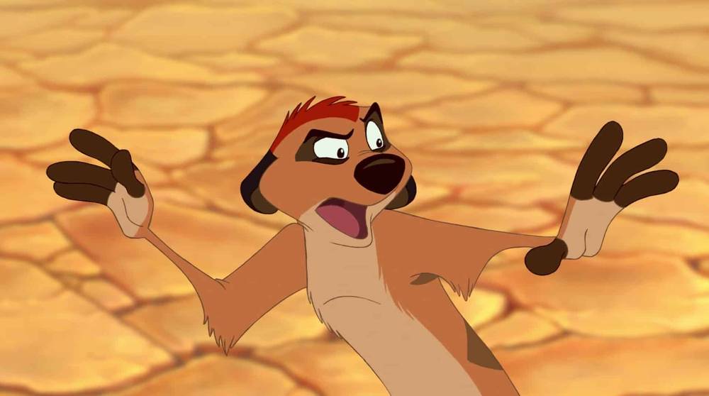 Image animée de Timon du Roi Lion de Disney, l'air surpris, la bouche ouverte et les bras tendus, sur un arrière-plan flou de terre sèche et craquelée.