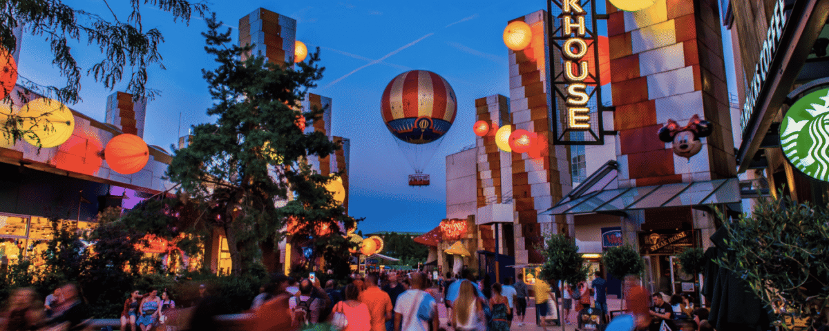 Scène nocturne animée à Disney Village, avec des lanternes illuminées, une publicité pour des montgolfières et des passants passant devant divers restaurants et magasins.