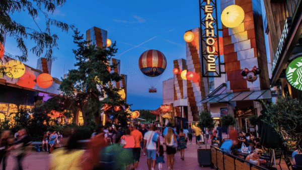 Scène nocturne animée à Disney Village, avec des lanternes illuminées, une publicité pour des montgolfières et des passants passant devant divers restaurants et magasins.