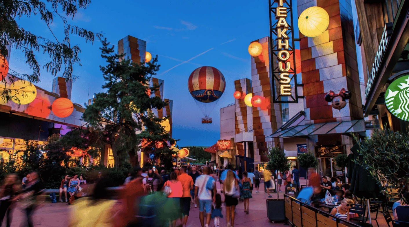 Scène nocturne animée à Disney Village, avec des lanternes illuminées, une publicité pour des montgolfières et des passants passant devant divers restaurants et magasins.