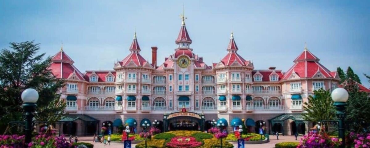 Une vue pittoresque du Newport Bay Club avec une architecture rose vif, avec des tours ornées et une horloge, entourée d'une verdure luxuriante et de fleurs colorées sous un ciel nuageux.