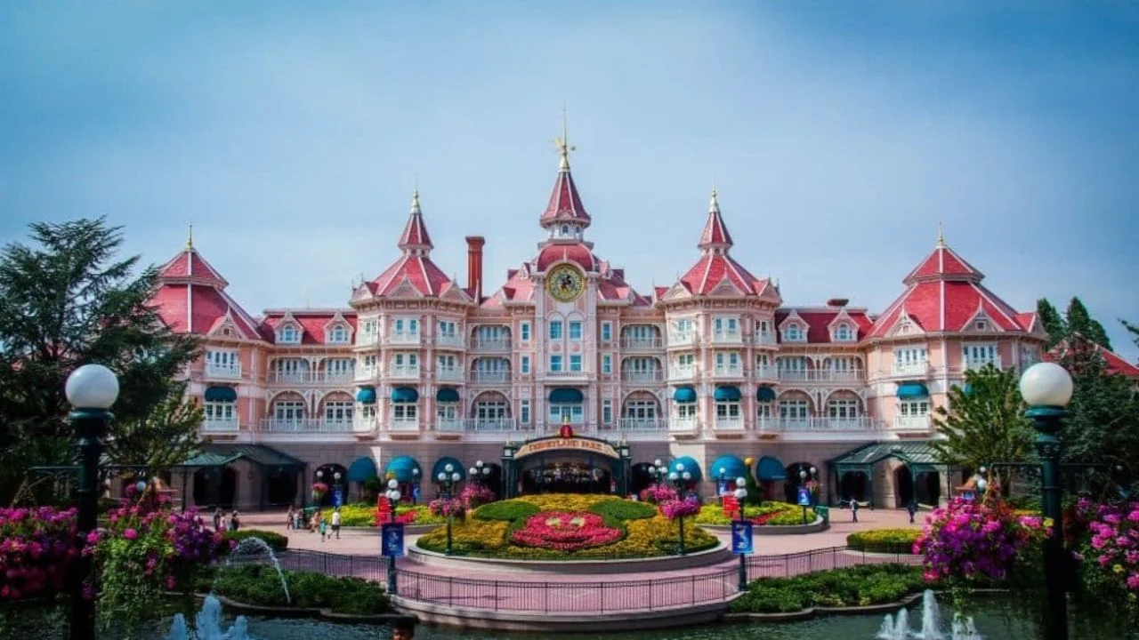Une vue pittoresque du Newport Bay Club avec une architecture rose vif, avec des tours ornées et une horloge, entourée d'une verdure luxuriante et de fleurs colorées sous un ciel nuageux.