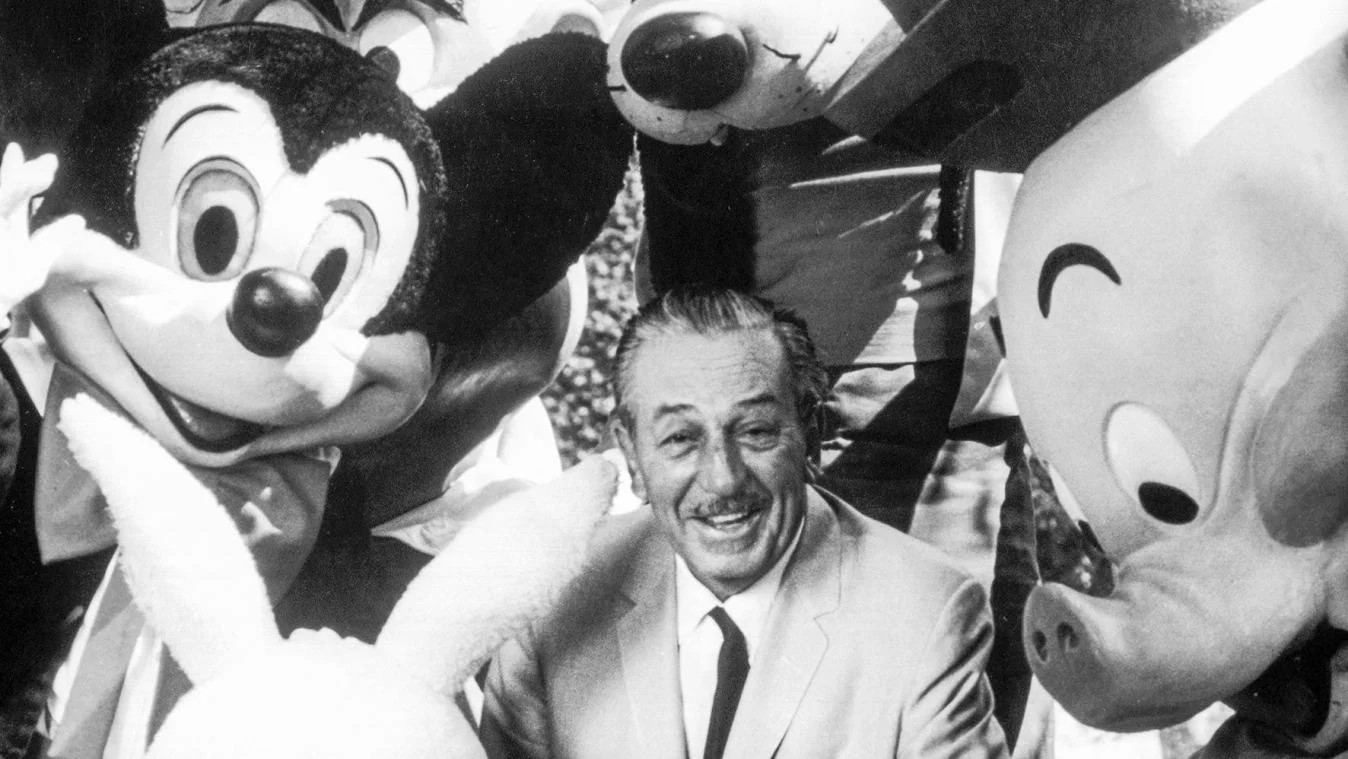 Walt Disney sourit avec Mickey Mouse et Porky Pig lors de la journée d'ouverture de Disneyland en 1955, célébrant la magie de l'animation.