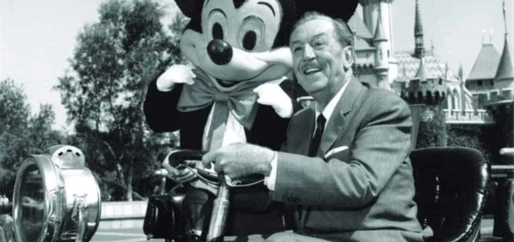 Walt Disney assis sur une grue-caméra vintage avec une grande figurine de Mickey Mouse, souriante, avec en arrière-plan le Château de la Belle au Bois Dormant de Disneyland, emblématique de l'industrie du divertissement.
