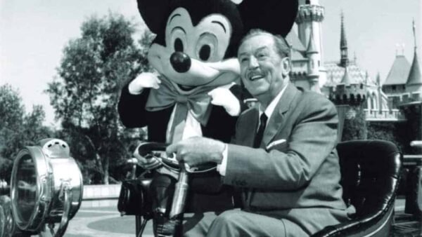 Walt Disney assis sur une grue-caméra vintage avec une grande figurine de Mickey Mouse, souriante, avec en arrière-plan le Château de la Belle au Bois Dormant de Disneyland, emblématique de l'industrie du divertissement.
