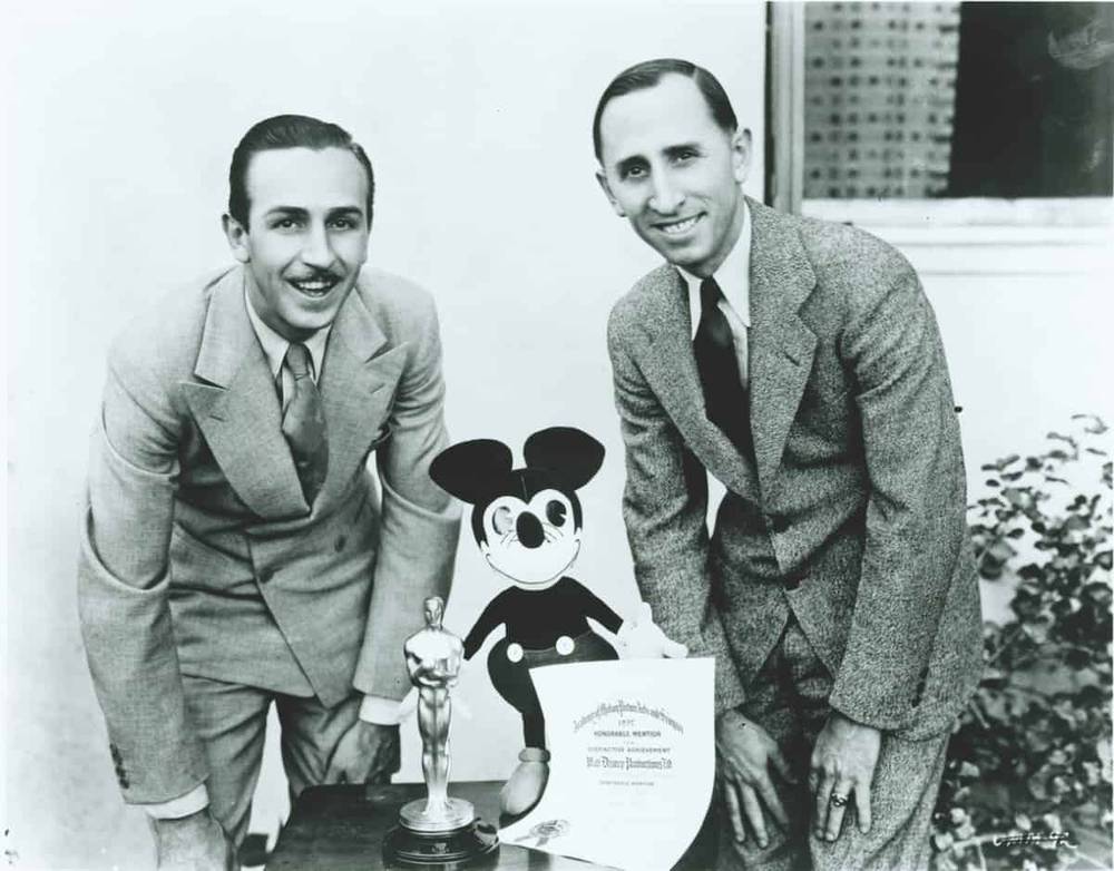 Walt Disney et Ub Iwerks posant avec une illustration de Mickey Mouse, un Oscar et un certificat. Ils sont habillés en costumes, souriants, sur fond urbain dans le monde de l'animation